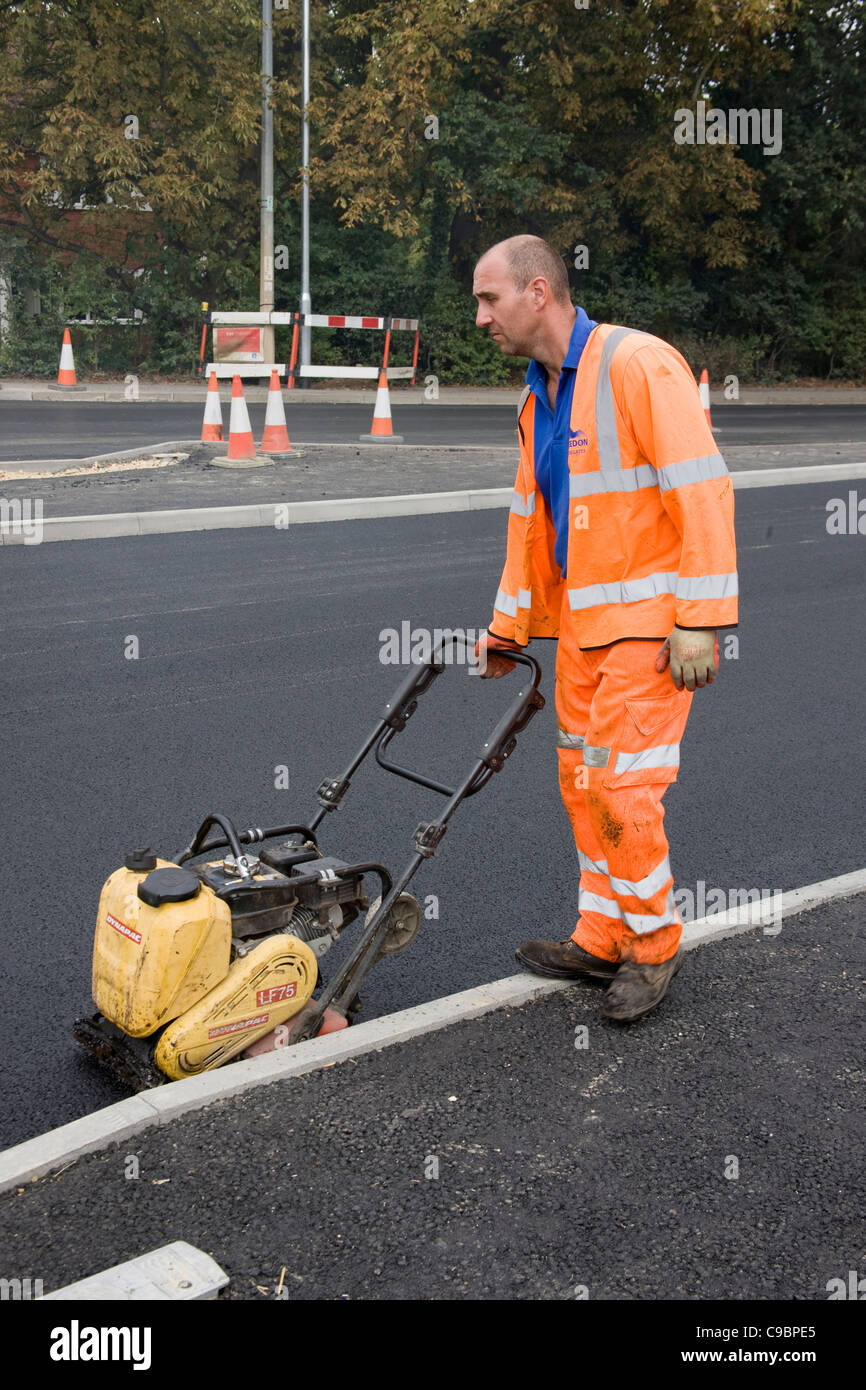 Workman utilizzando una dynapac per compact asfalto (asfalto) lungo il bordo del marciapiede (cordolo) Foto Stock