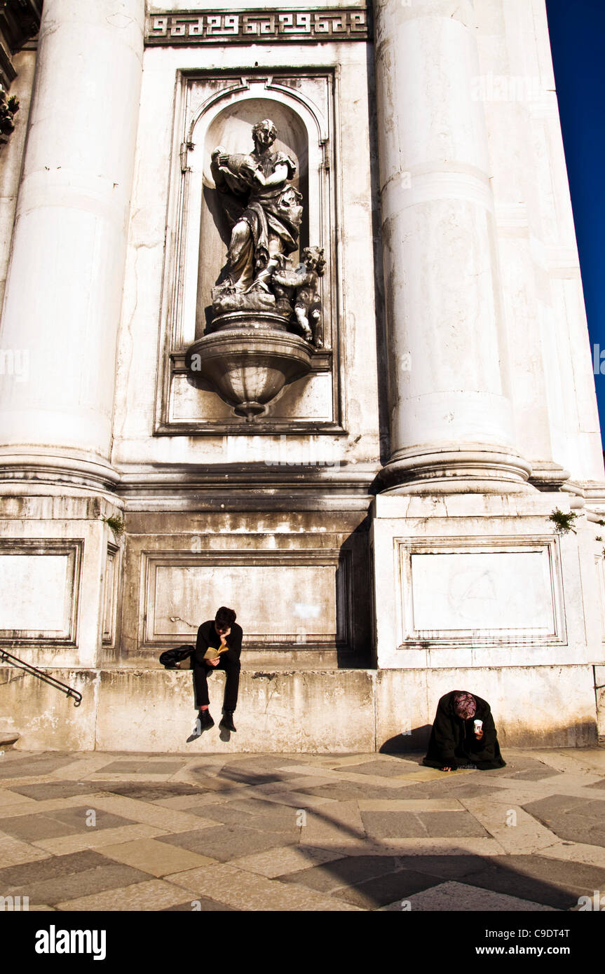 Venezia Italia. Una donna sulle sue ginocchia mendica per denaro come un giovane uomo legge un libro al di fuori della chiesa di Santa Maria del Rosario sul Canale della Giudecca Foto Stock