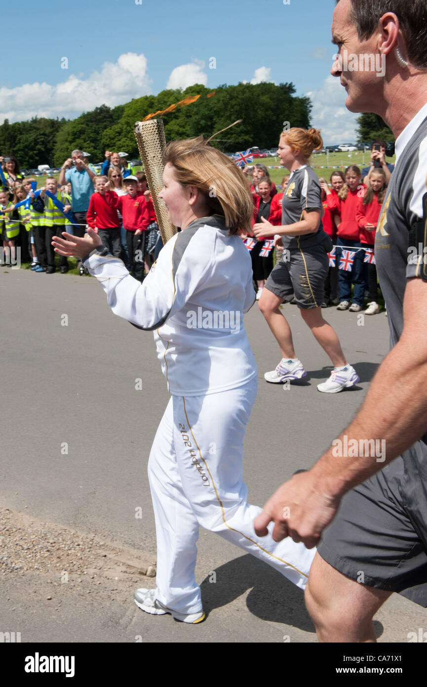 Olympic Torchbearer Janet Baker (portante & holding torce fiammeggianti aloft) è guardata da grande folla di ben wishers & accompagnata dalla torcia di sicurezza i membri del team in grigio kit in esecuzione. Nella motivazione della Harewood House nello Yorkshire, Inghilterra, Regno Unito. Martedì 19 Giugno 2012. Foto Stock