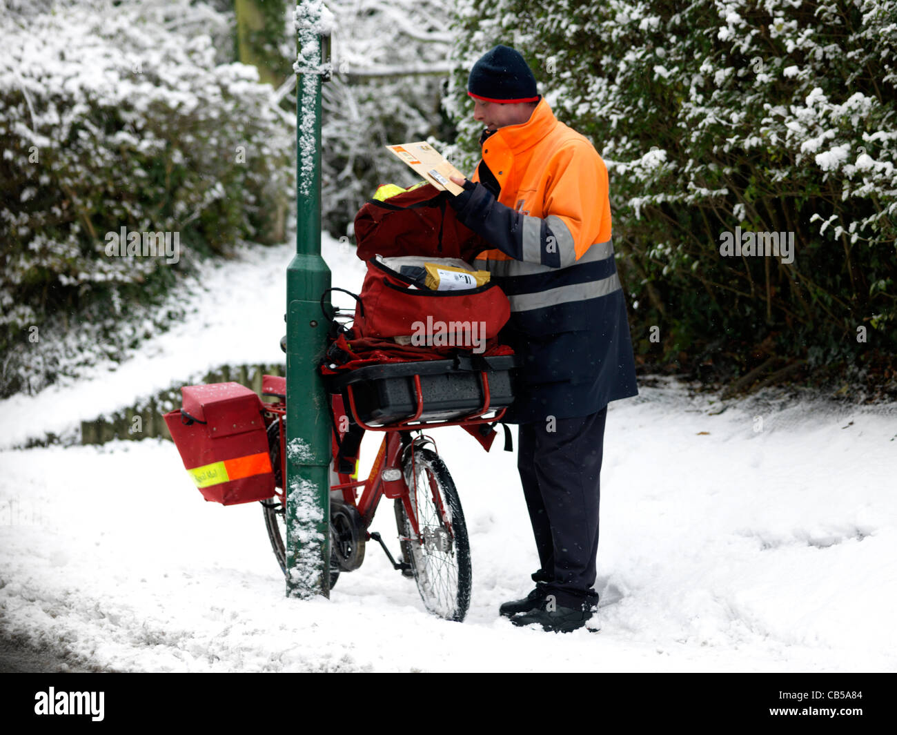 Portalettere Sorting Out Post borsa con la bicicletta appoggiata contro la lampada posta nella neve Surrey in Inghilterra Foto Stock