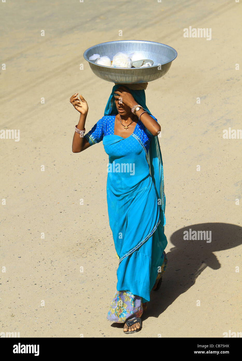 Donna indiana in abito tradizionale su una strada polverosa che porta il suo cibo sul suo capo. Il Rajasthan. India Foto Stock