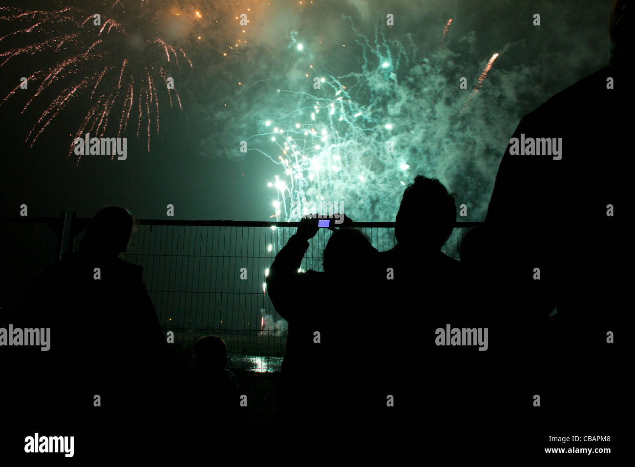 I fuochi d'artificio in London, luminosi di colore verde e rosso esplosioni riempiono il cielo mentre si stagliano le persone guardano da dietro una recinzione Foto Stock