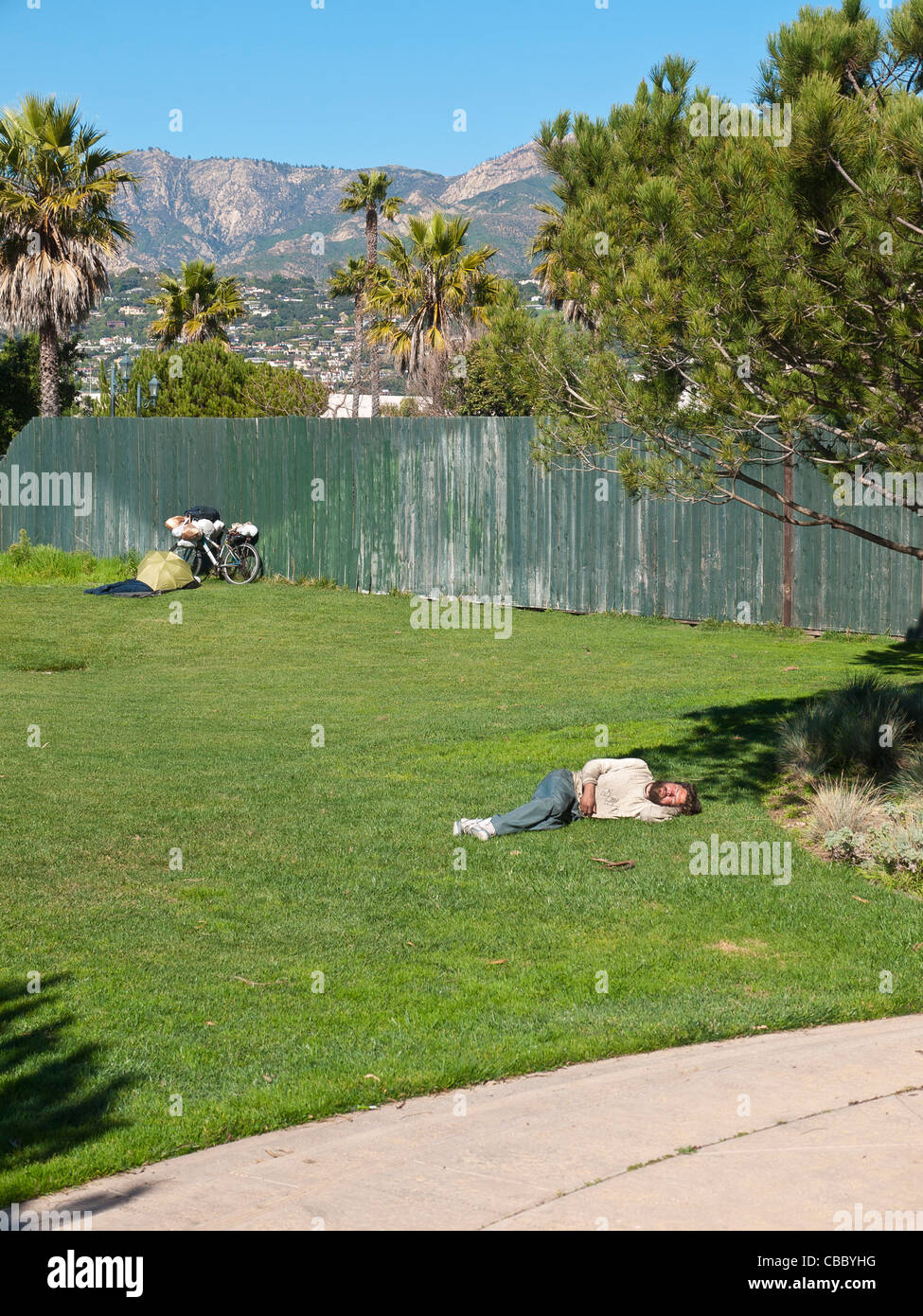 Una trentina di qualcosa di senzatetto uomo dorme mezzogiorno sul prato verde di un parco della città con la sua bicicletta. Foto Stock