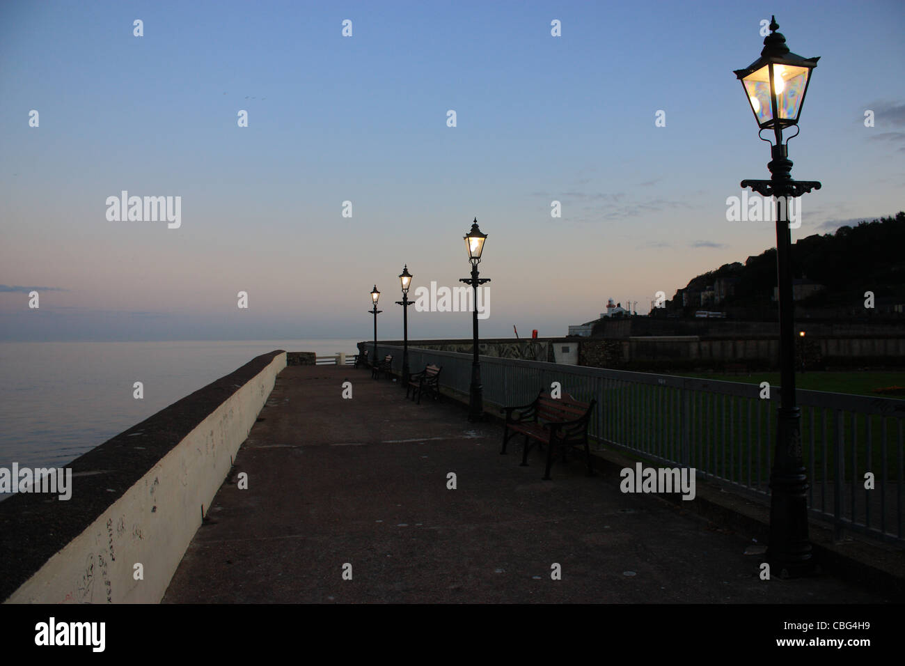 Un molo al tramonto sulla costa meridionale dell'Irlanda. lampade illuminate stare accanto a un mare calmo con un faro lontano in background Foto Stock