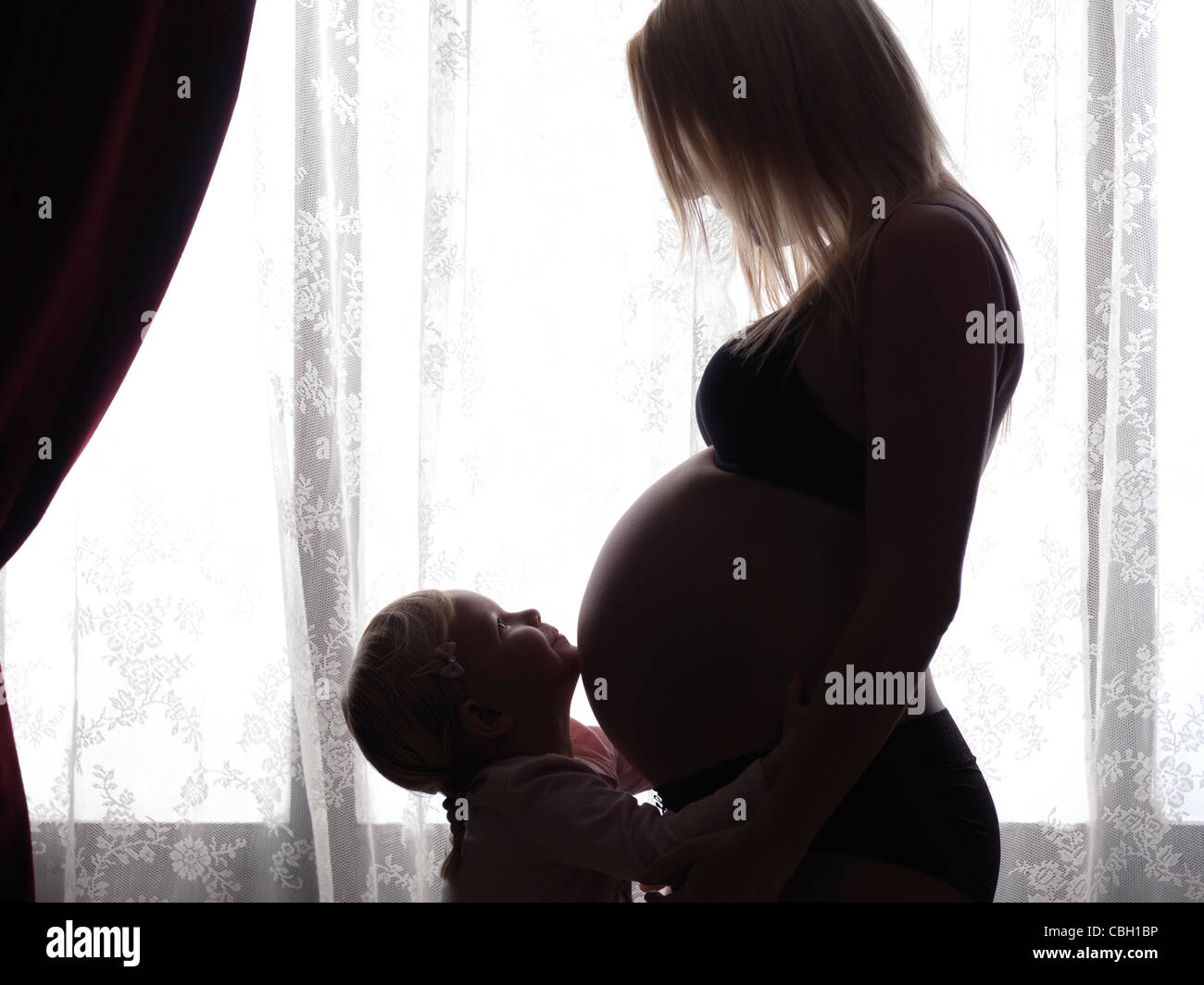 Felice piccola figlia abbracciando la sua gravidanza madre della pancia in luce davanti a una finestra Foto Stock
