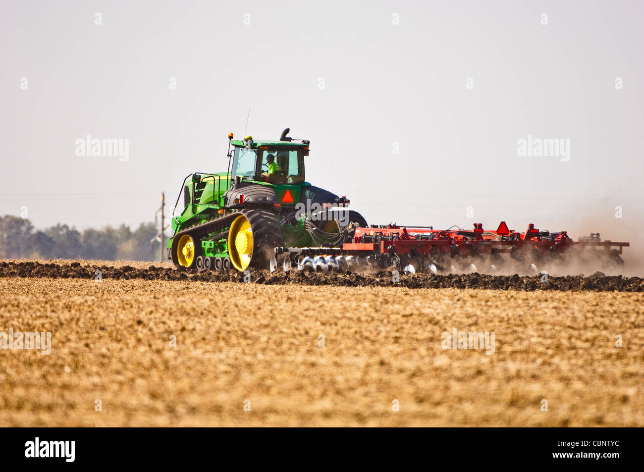 Un moderno trattore cingolato aratri un campo dopo il raccolto. Alta venti soffiano il secco e sfuso terreno superiore come l'aratro rompe la terra. Foto Stock
