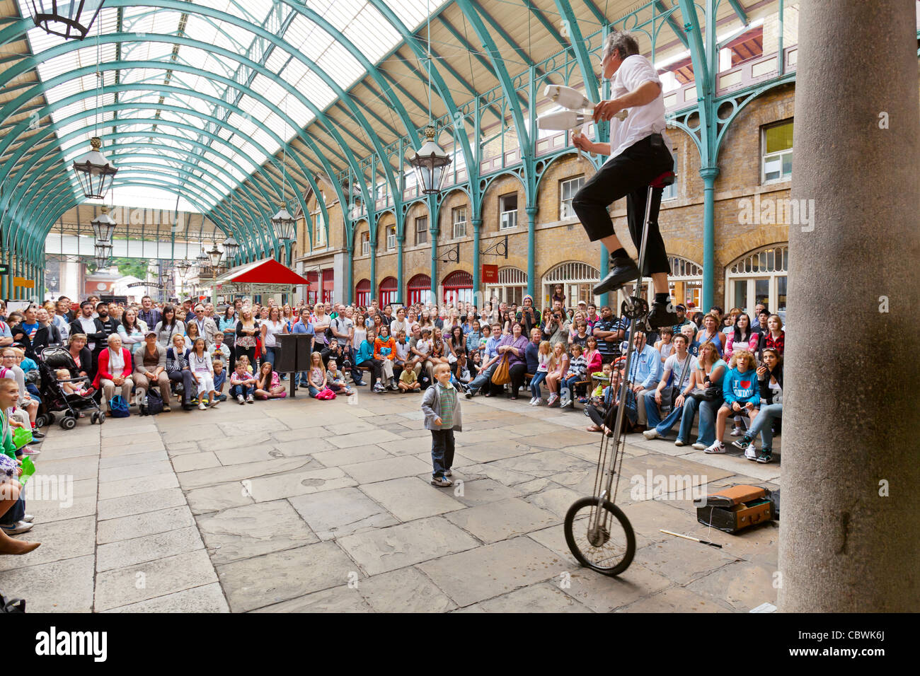 I turisti e gli amanti dello shopping e un attore di strada su una bicicletta alto con un pubblico al Covent Garden di Londra, Inghilterra. Foto Stock