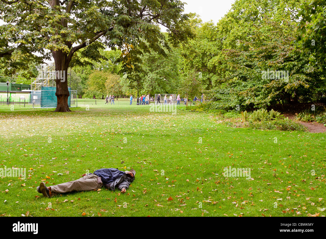 Senzatetto uomo dorme in St James Park, Londra, Inghilterra mentre la gente sta giocando il gioco della palla in background. Foto Stock