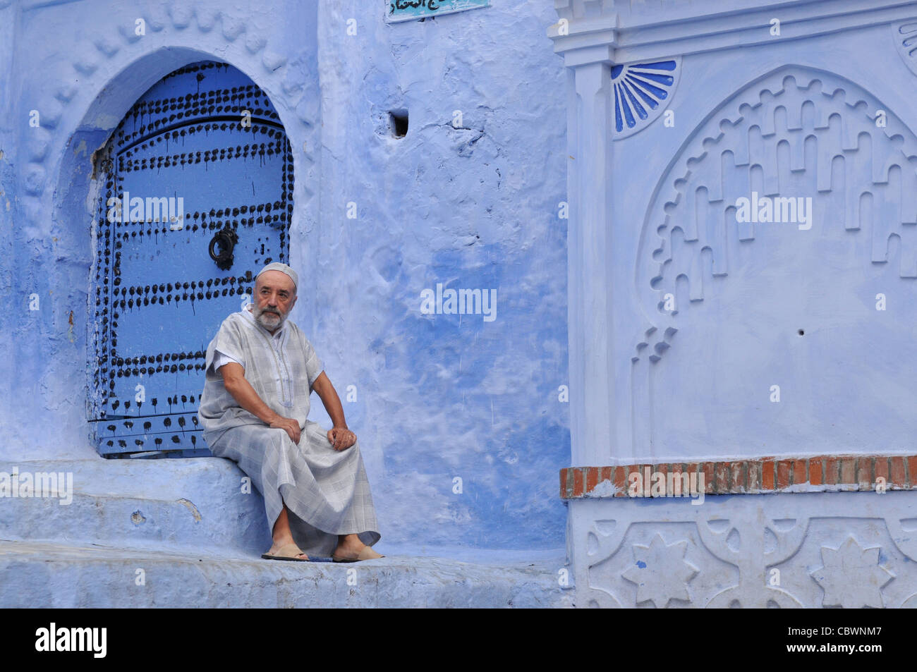 Tradizionalmente vestito uomo appoggiato vicino blu lavato case nelle strade di Chefchaouen Marocco Africa del Nord Foto Stock