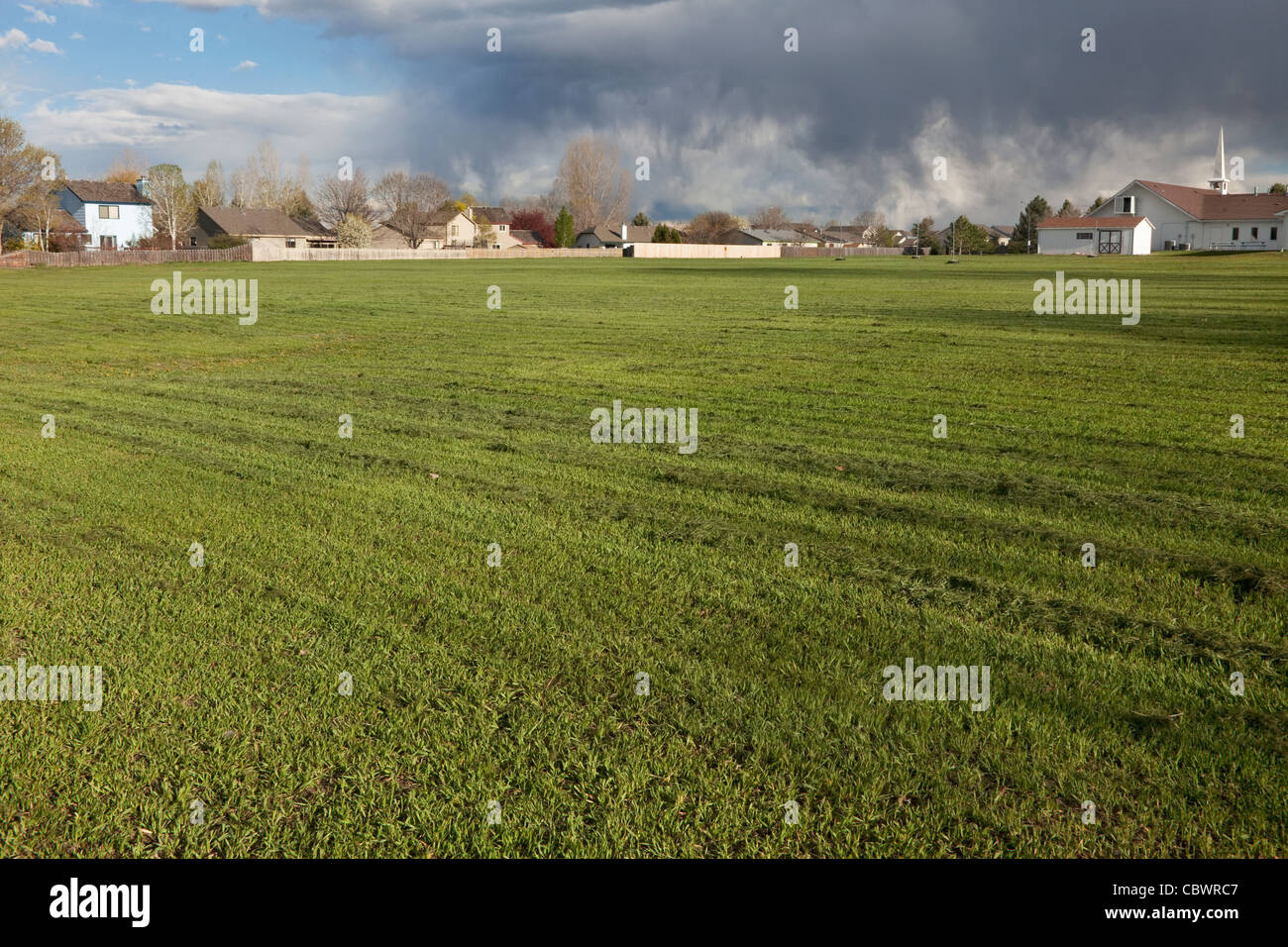 Appena falciata, ampio campo di erba con case residenziali, chiesa e pesanti nuvole temporalesche in background Foto Stock