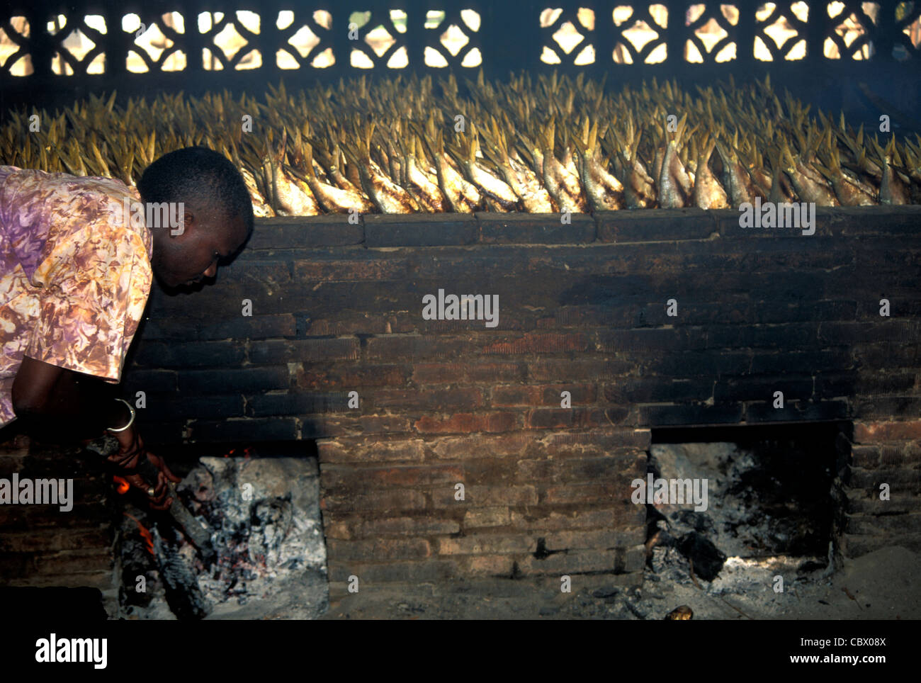 Un uomo che alimenta il fuoco in una casa di fumo di pesce in Gambia, Africa occidentale Foto Stock