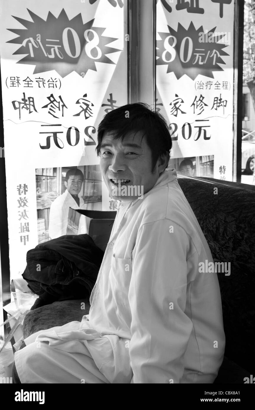 Pechino. Il sig. Bao LinDi, un tradizionale pedicurist, seduti dietro il suo negozio di finestra. Foto Stock