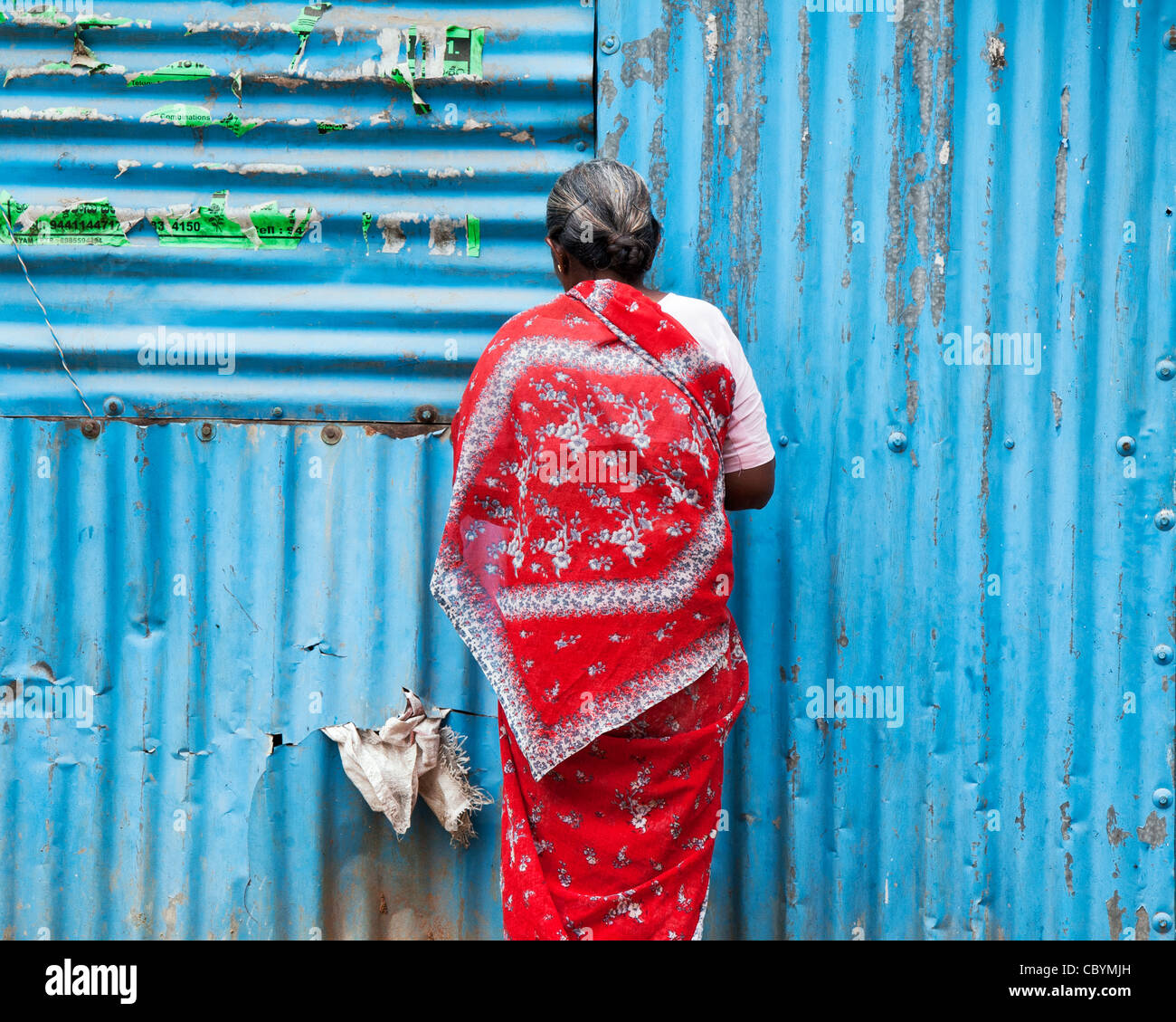 Il vecchio donna indiana in un sari rosso in piedi di fronte ad uno stagno blu shack. Andhra Pradesh, India Foto Stock