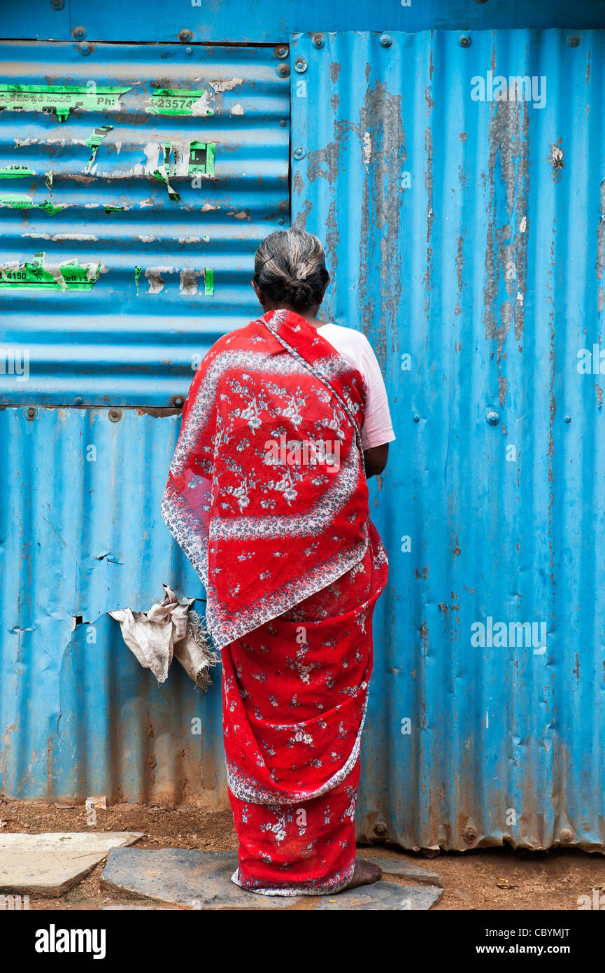 Il vecchio donna indiana in un sari rosso in piedi di fronte ad uno stagno blu shack. Andhra Pradesh, India Foto Stock