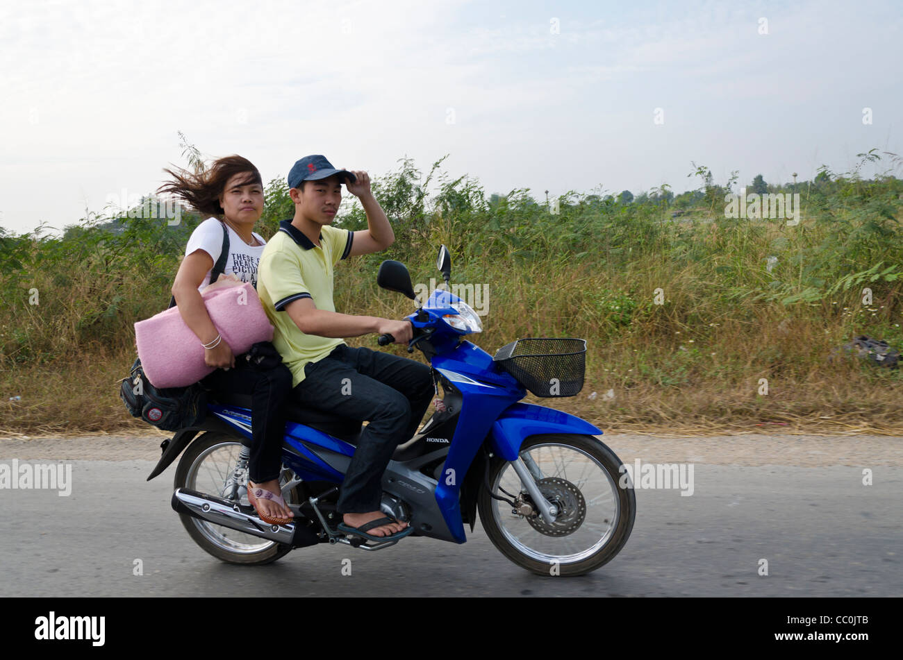 Giovane uomo Thai & donna che viaggia sul motociclo blu guardare fotocamera. L uomo sta tenendo in mano il suo cappello & donna è in possesso di una coperta ripiegata. Foto Stock