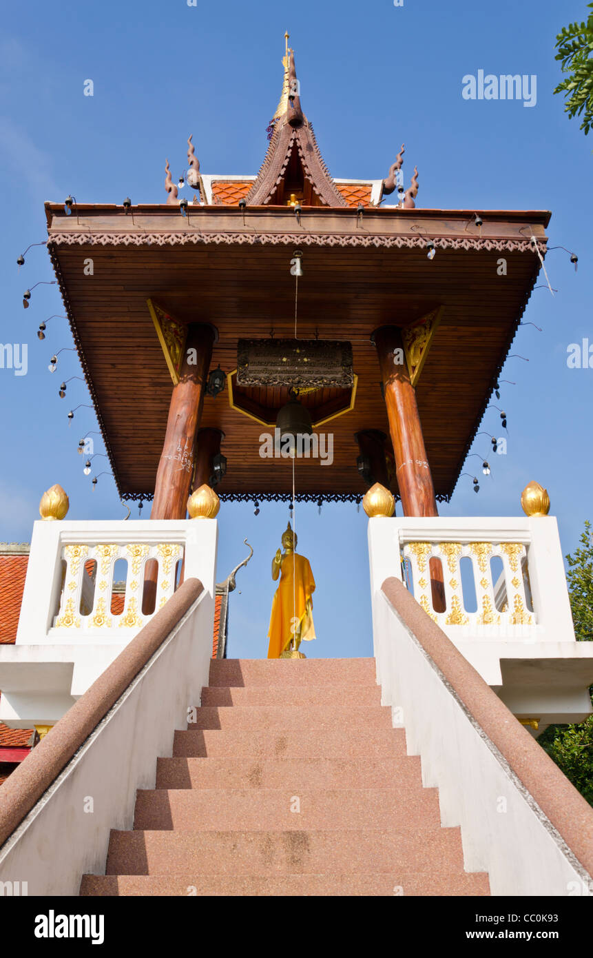Una scalinata conduce fino a oro statua del Buddha con scialle drappeggiati su di esso & campana grande sopra di esso al tempio buddista in Thailandia del Nord Foto Stock