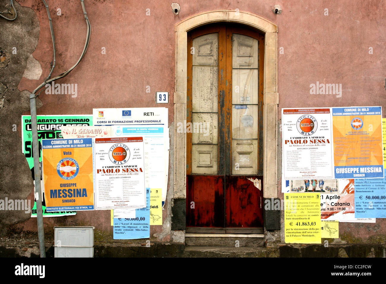 Una piccola porta con il numero 93 accanto ad essa contro una parete rossa in Sicilia, Italia. Poster per feste locali decorano le pareti. Foto Stock