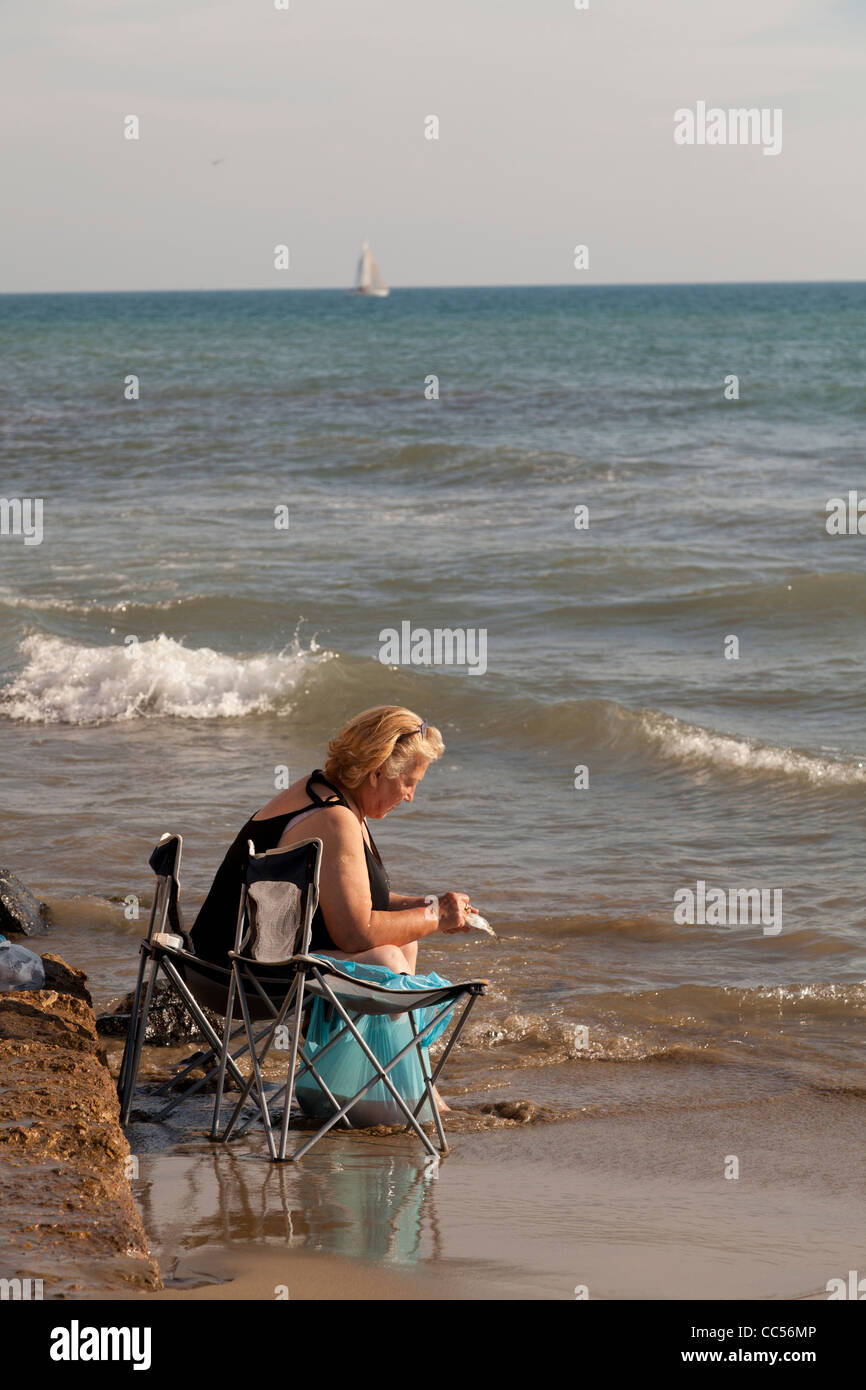 La donna sedeva al bordo delle acque della spiaggia di pesce di pulizia Foto Stock