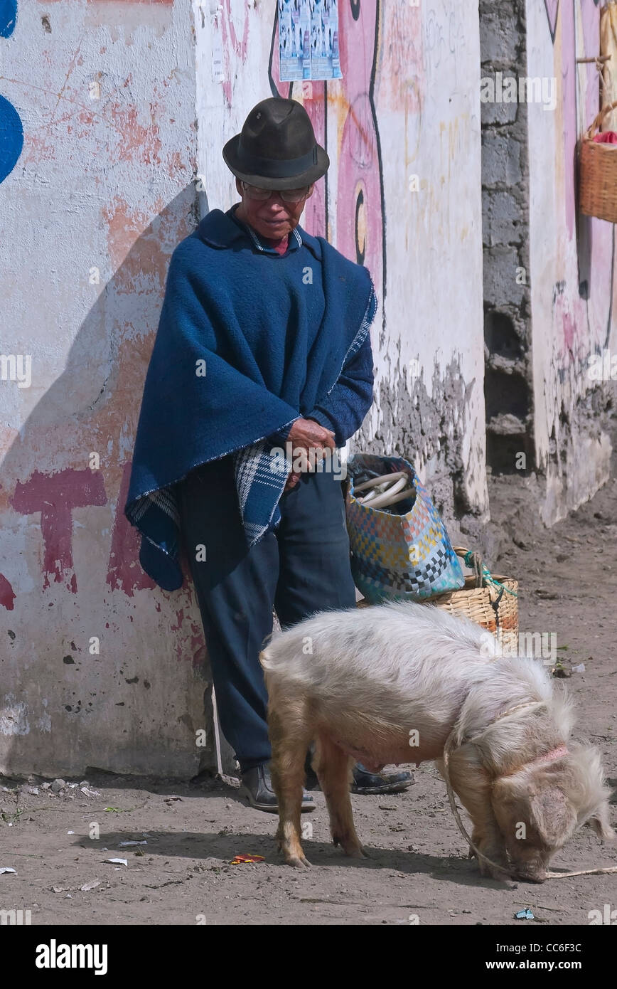 Un uomo di mezza età in tradizionale abbigliamento ecuadoriana si appoggia contro lo spigolo di un edificio con un maiale al guinzaglio davanti a lui. Foto Stock