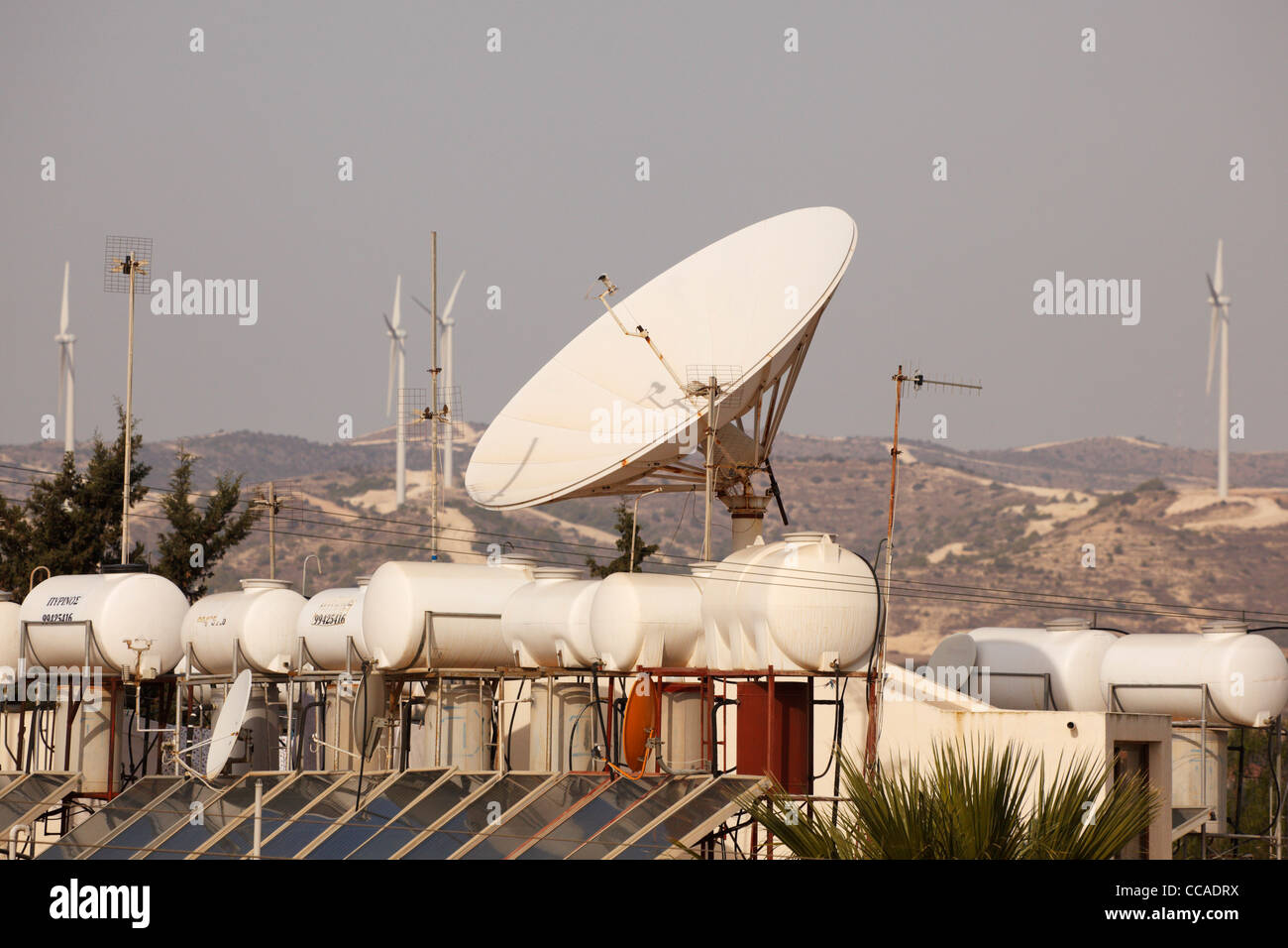 Pannelli solari e serbatoi, grande parabola satellitare sul tetto con turbine eoliche dietro. Pervolia, Cipro Foto Stock