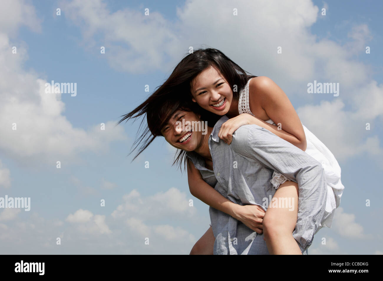 Giovane uomo che porta una donna sulla sua schiena ridendo, cielo blu con nuvole come sfondo Foto Stock
