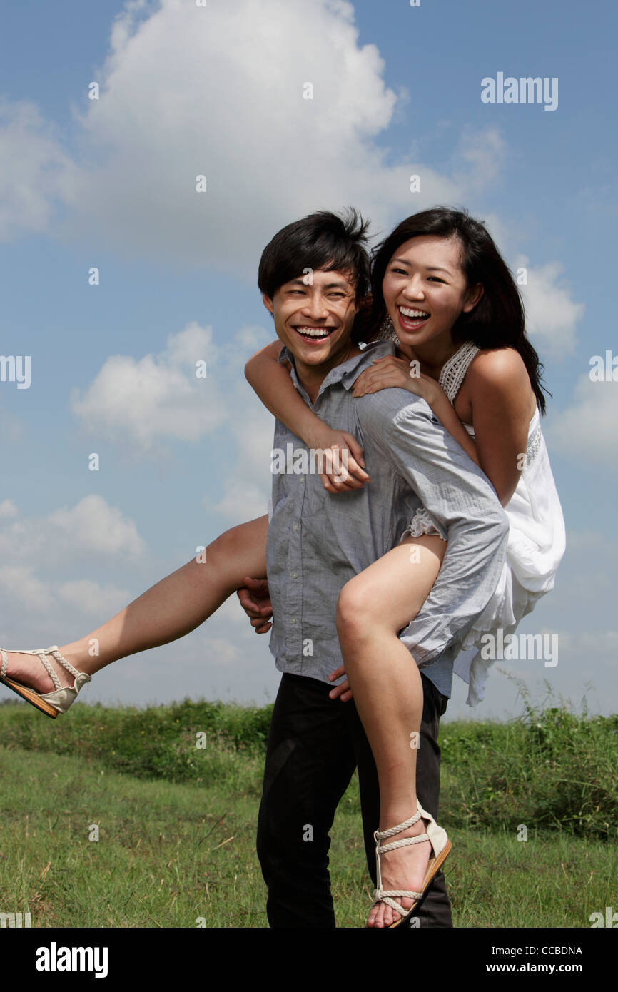 Giovane uomo che porta una donna per il suo ritorno a ridere con cielo blu e nuvole in background. Foto Stock