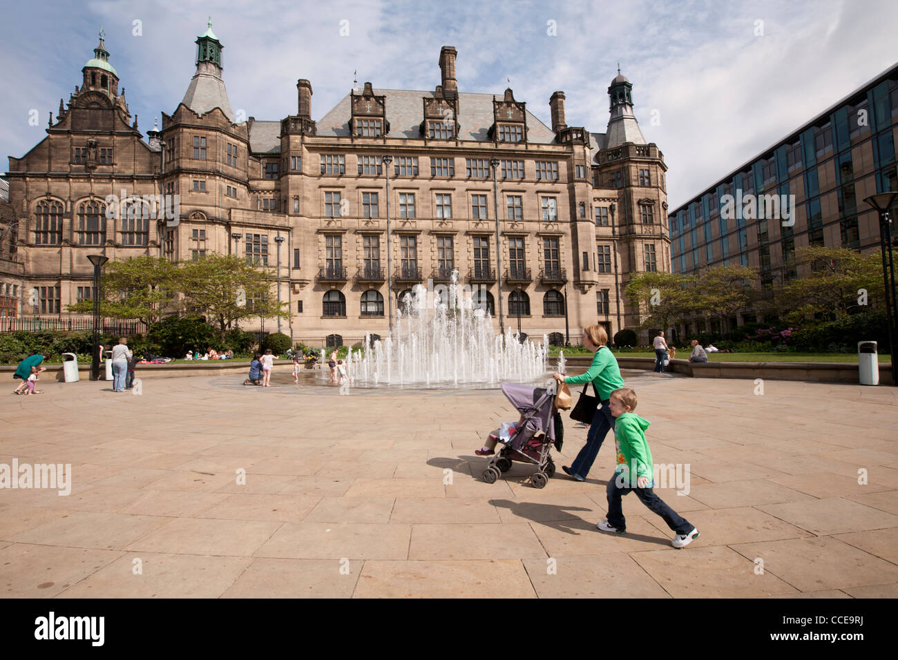 Una madre e un bambino sia vestito in verde le felpe, madre con un buggy, a piedi di fronte alla fontana e Sheffield Town Hall Foto Stock