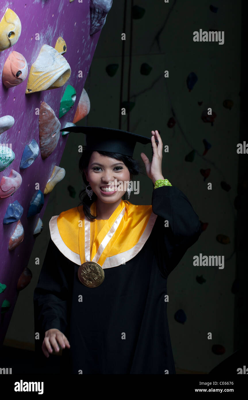 Giovane attraente Asian Donna recentemente laureando roccia arrampicata su una parete artificiale arrampicata per rappresentare il successo Foto Stock