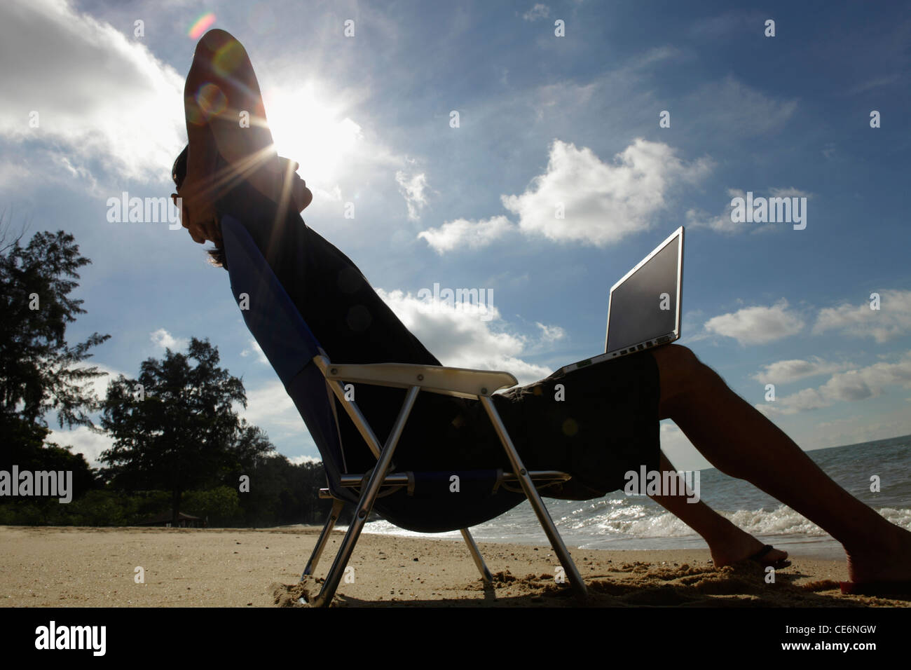 Uomo seduto nella sedia spiaggia con laptop, sun burst dietro la testa Foto Stock