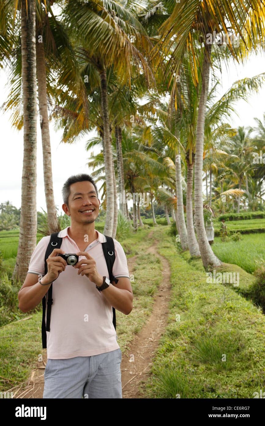 Uomo in piedi su un piccolo sentiero tenendo una fotocamera nella parte anteriore degli alberi di cocco. Foto Stock