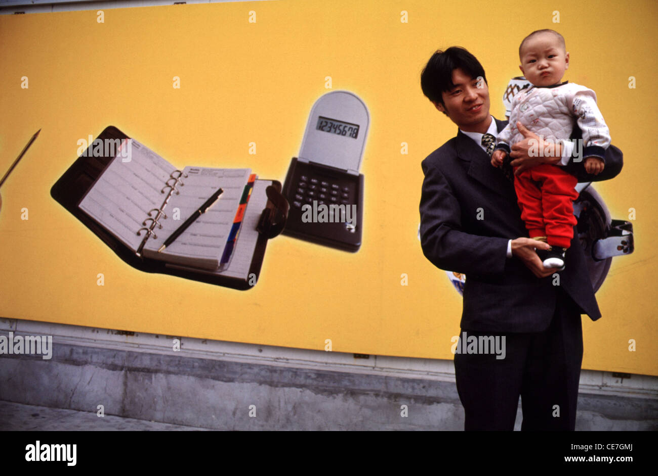 Un giovane uomo cinese in abito nero tiene il suo figlio davanti a un poster con apparecchi per ufficio in Cina Foto Stock