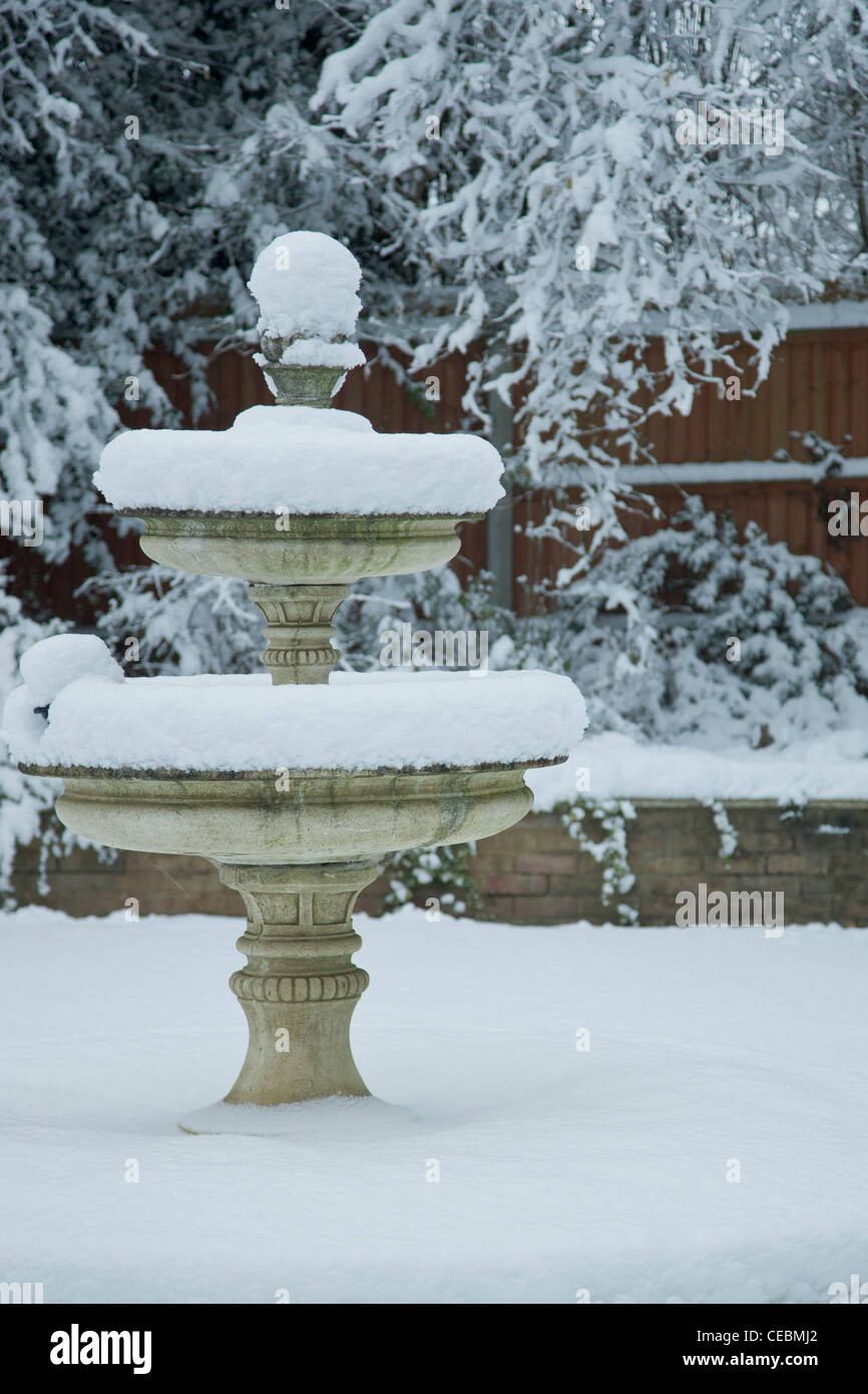 Una fontana sorge impilati con neve su ciascuno dei suoi tre livelli con piante e una recinzione in background Foto Stock