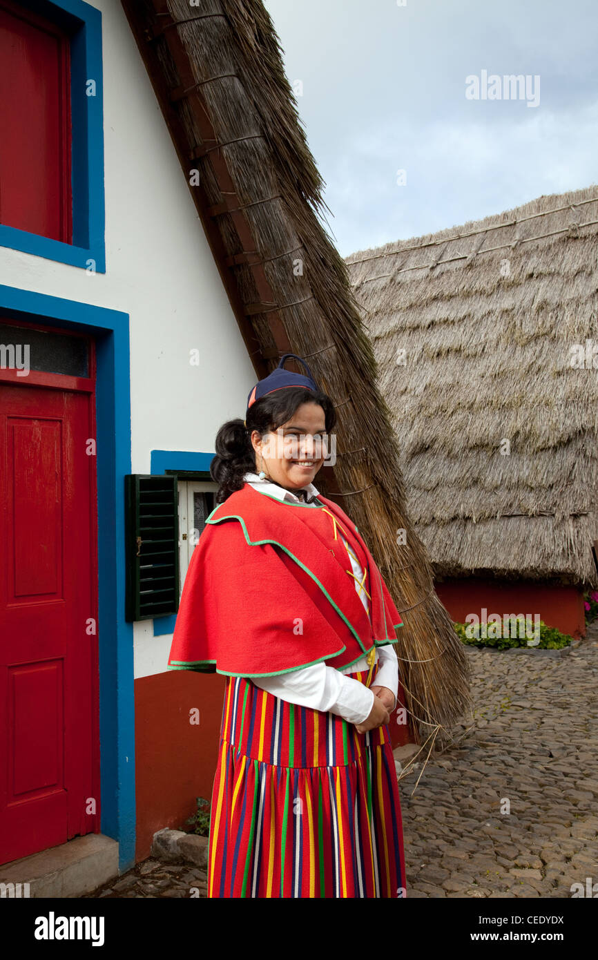 Tradizionale casa portoghese Palheiro con struttura triangolare e tetto in paglia. Donna vestita in costume tradizionale con tessuti multicolore Santana, Madeira Foto Stock