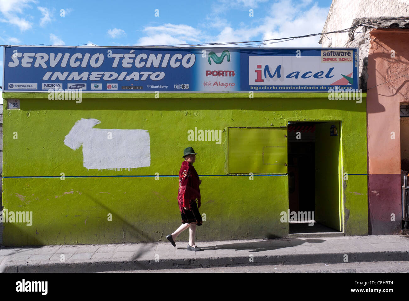 Una donna indigena passeggiate da una chartreuse edificio colorato che è un telefono cellulare repair shop strada laterale in Latacunga, Ecuador. Foto Stock