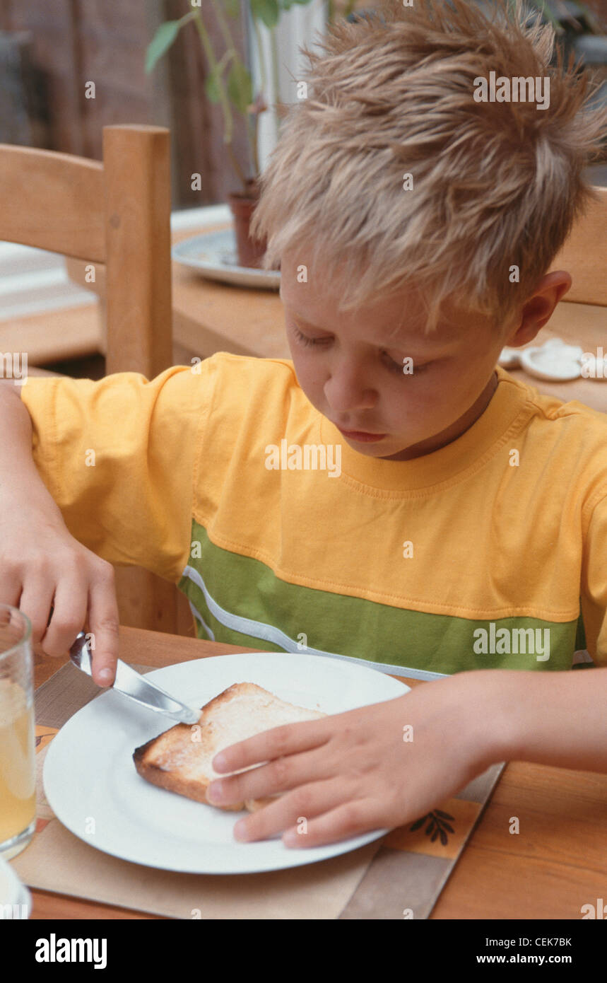Bambino maschio pungenti capelli biondi che indossa il verde e il giallo t shirt seduti al tavolo di legno guardando giù diffusione di burro sul pane tostato Foto Stock