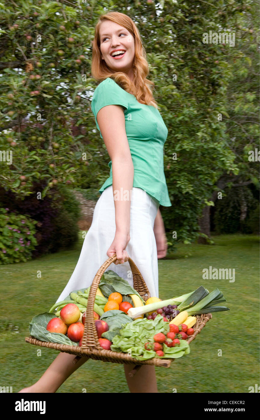 Luce femmina marrone capelli indossando un verde di T-shirt e un mantello bianco, portando un cesto di giardinaggio di frutta e verdura cercando Foto Stock