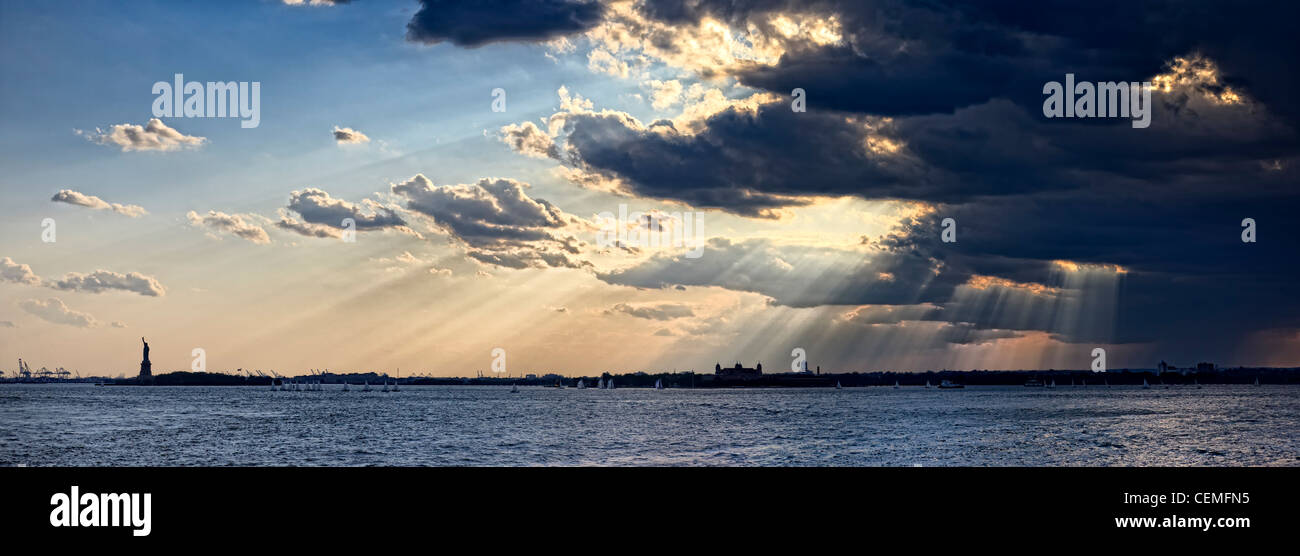 Golden raggi di sole piercing attraverso le nuvole scure, e Shining sulla Statua della Libertà e Ellis Island del porto di New York, Stati Uniti d'America Foto Stock