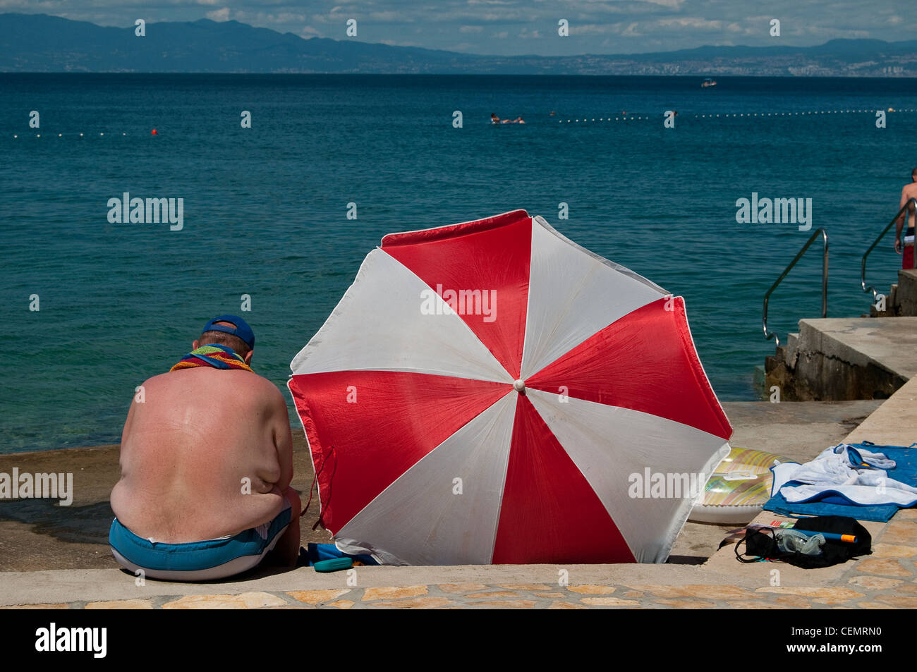 L'uomo siede sull'acqua su una spiaggia di Croazia accanto al suo ombrello rosso e bianco. Foto Stock