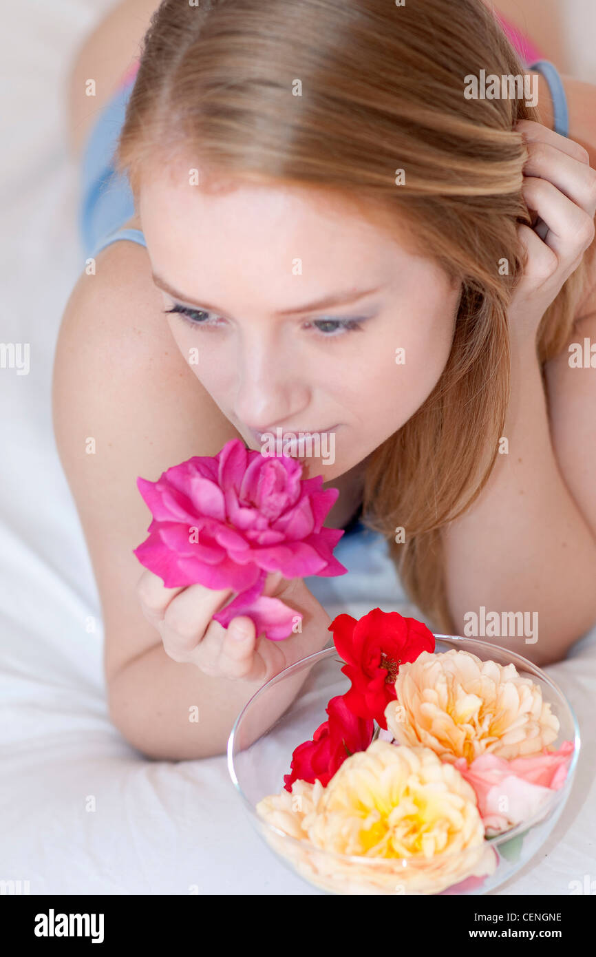 Femmina dritti lunghi capelli biondi, giacente sul letto sulla sua parte anteriore di una ciotola di vetro colorato e roses nella parte anteriore del suo profumo di rosa Foto Stock