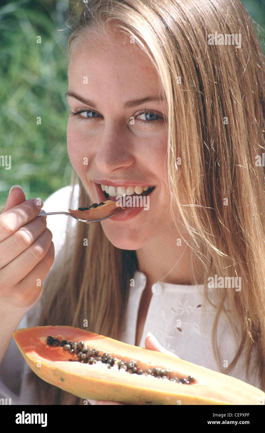 Femmina fiera lunghi capelli indossare top bianco broderie anglaise dettaglio, mangiando papaia cucchiaio cercando di fotocamera a sorridere mostrando i denti Foto Stock