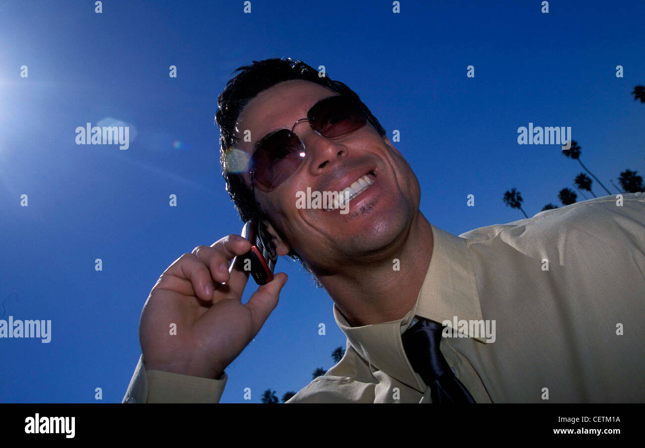 Maschio corto marrone capelli piccola ciocca di capelli sotto il labbro inferiore indossando occhiali da sole giallo shirt tirante grigio telefono cellulare all'orecchio cercando Foto Stock