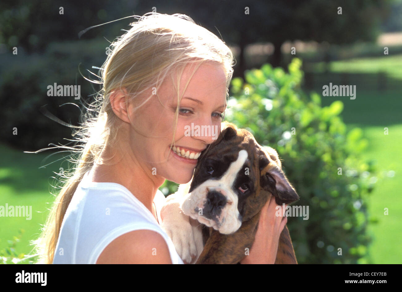 Felice femmina bionda cuddling suo marrone e bianco cane Boxer in un soleggiato parco Foto Stock