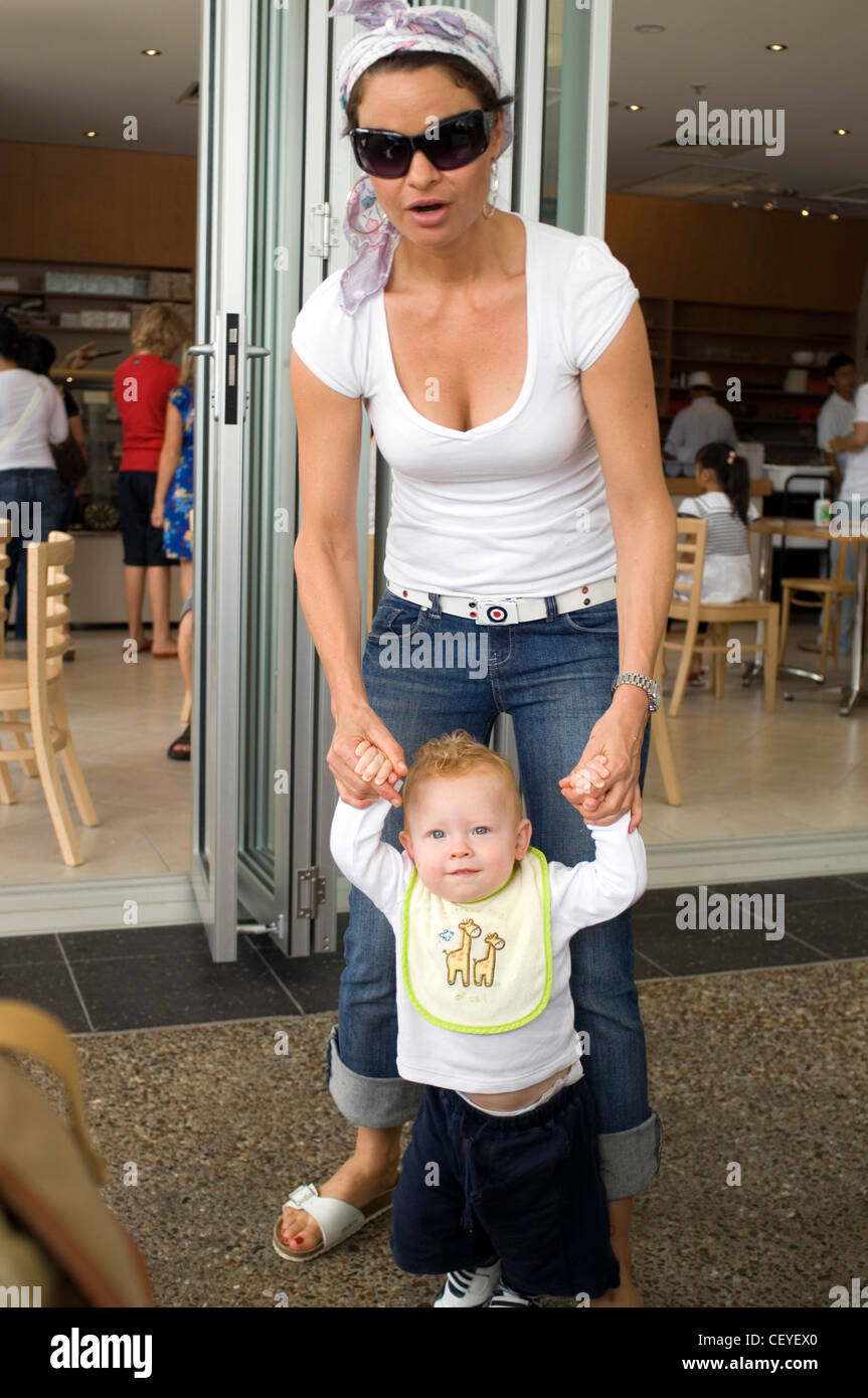 Femmina indossando occhiali da sole, testa sciarpa, bianco v di t-shirt, jeans cintura bianca e sandali bianchi, tenendo un biondo bambino maschio dal Foto Stock