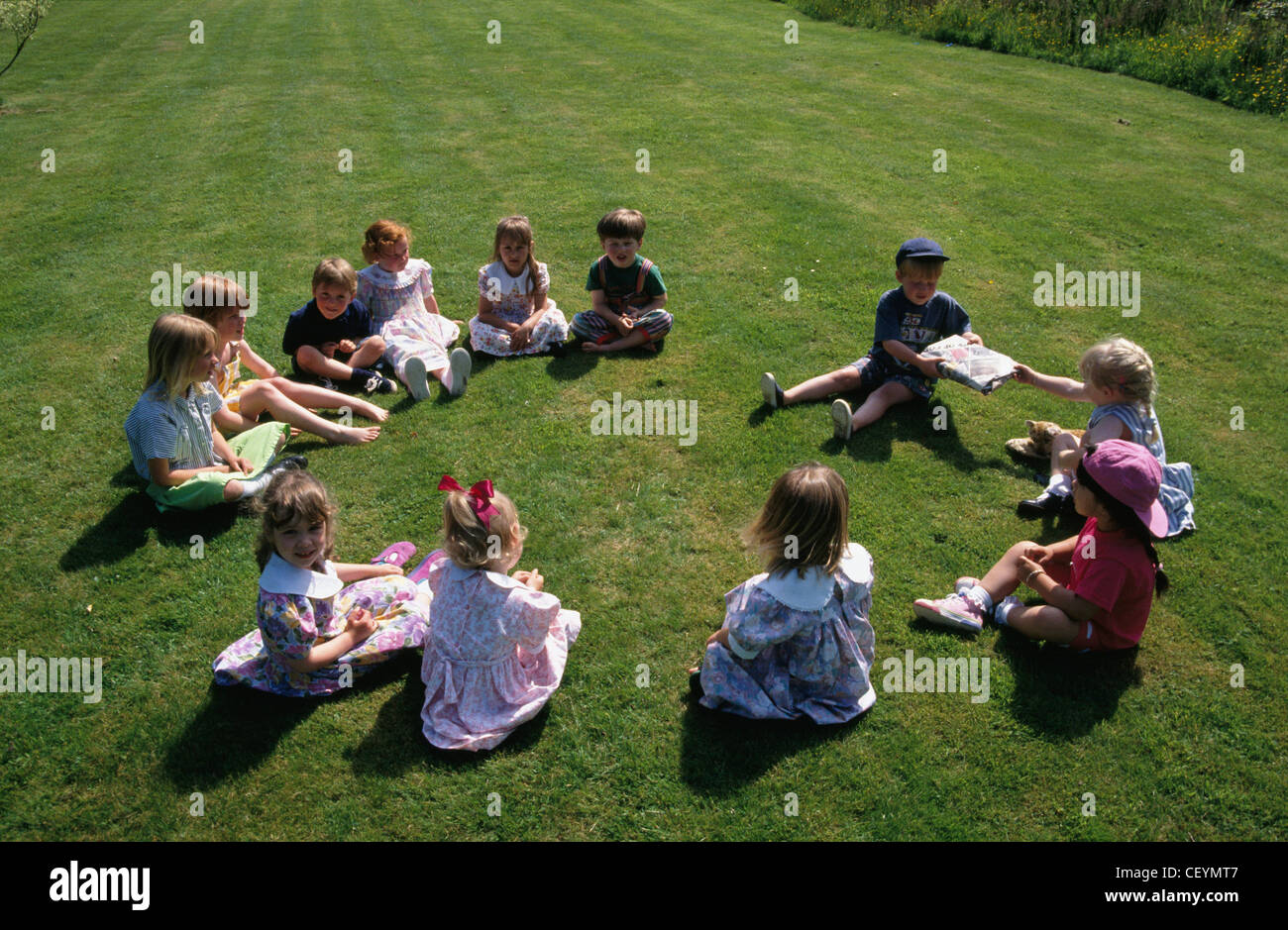 )Gruppo di bambini al di sotto di anni seduto sul prato la riproduzione di passare il pacco fotocamera premere Nicholas Holt Foto Stock