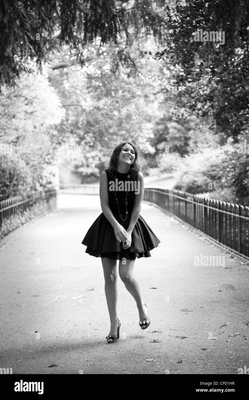 Donna che indossa un nero senza maniche abito flounced, collana lunga e scarpe dettagli di prua, in piedi nel parco, sorridente e i suoi occhi Foto Stock