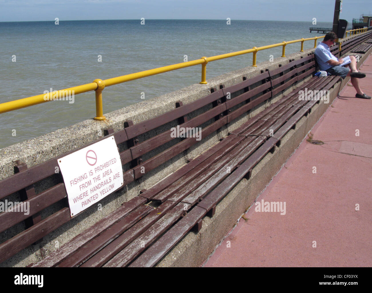 Un uomo legge un libro su un banco di lavoro sul molo ad affrontare, nel Kent, accanto a un corrimano giallo con nessun segno di pesca su di esso. Foto Stock