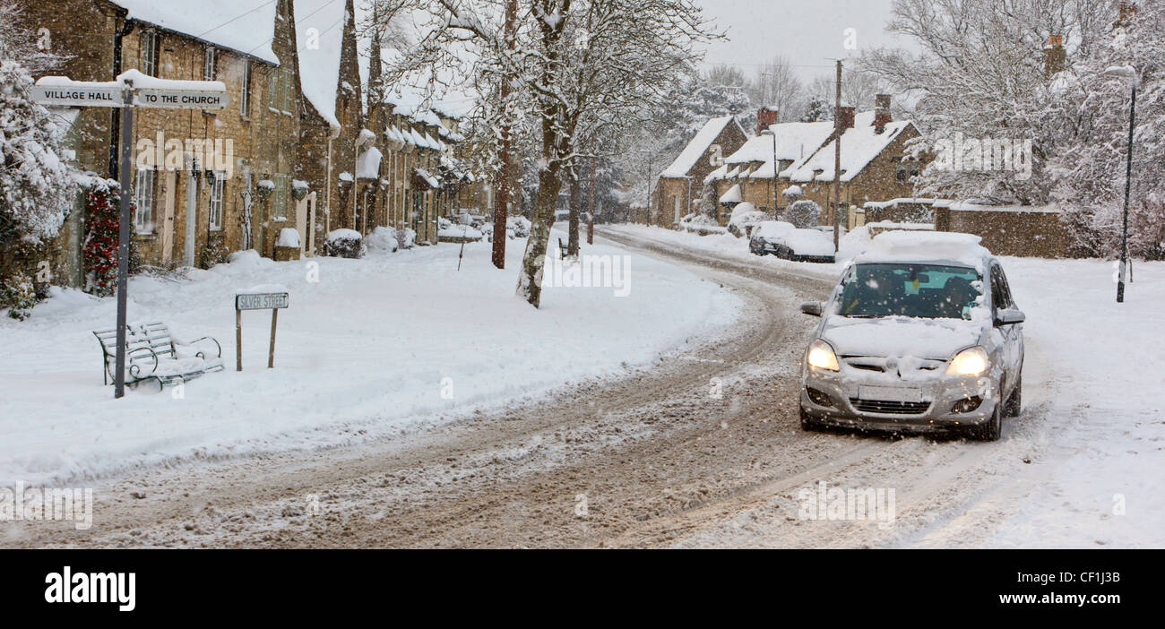 Una vettura lottando attraverso la caduta di neve sulla strada di argento in Cotswolds villaggio di South Cerney. Foto Stock