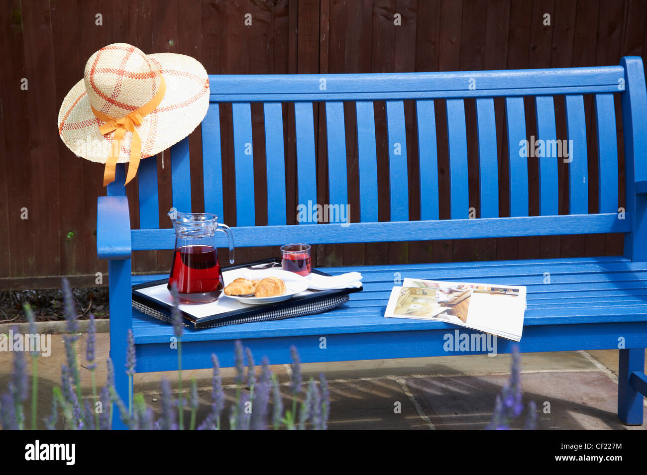Un dipinto di blu panchina da giardino, in un giorno d'estate, con il cappello  di paglia, la rivista e il vassoio di cui con drink di raffreddamento e  croissant Foto stock -