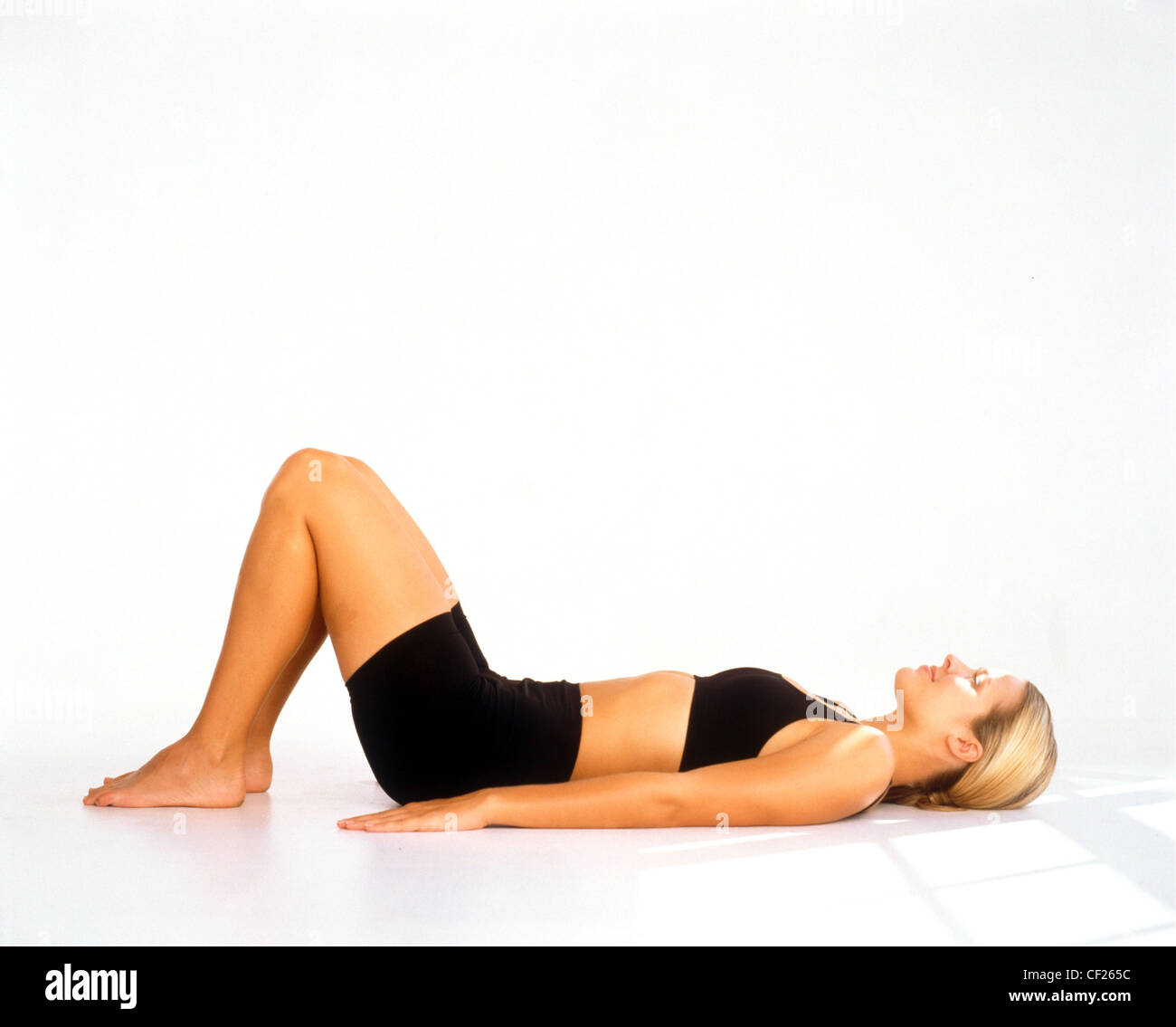 Profilo di donna capelli biondi vestita di nero reggiseno e shorts mentire indietro su floarms dal lato e ginocchia fino i piedi piatti sul pavimento Foto Stock