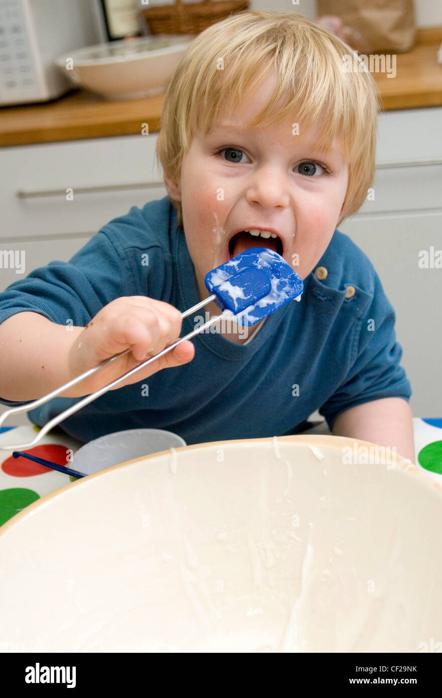 Un bambino maschio di mangiare torte al di fuori di un vaso che lambisce la spatola Foto Stock